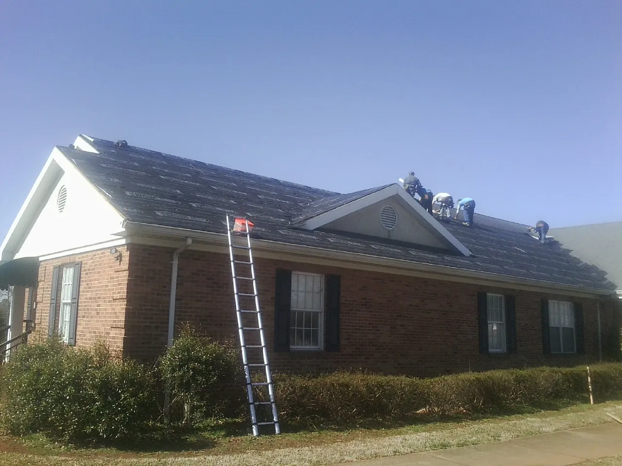Crew tearing shingles off a brick ranch roof with a ladder set — Metal Roof Repair work by our crew in Vance