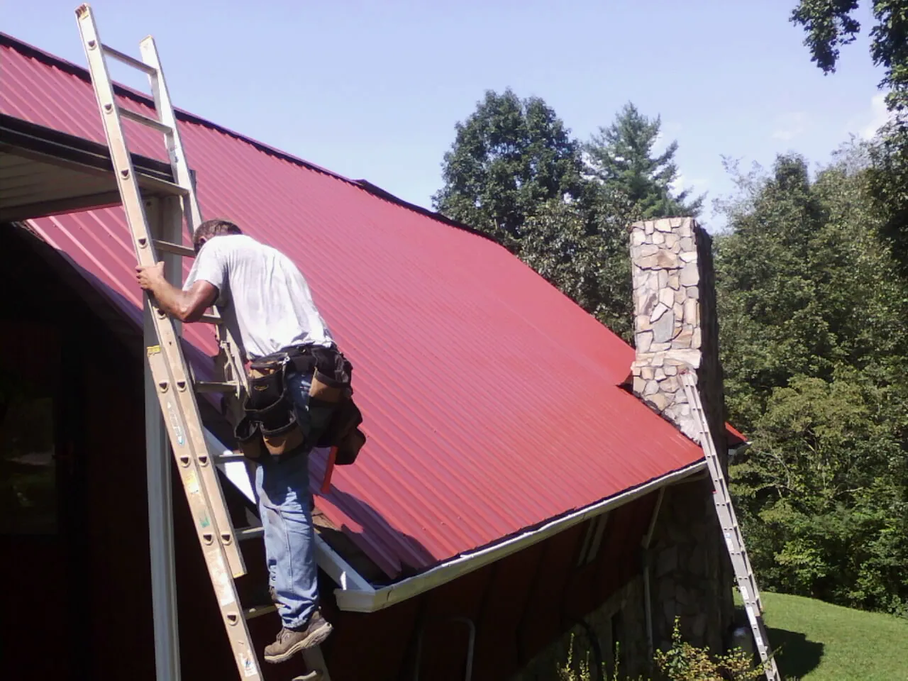 Roofer on a ladder beside a red metal roof and stone chimney — Soffit & Fascia Repair by Roofing Brothers in Vance